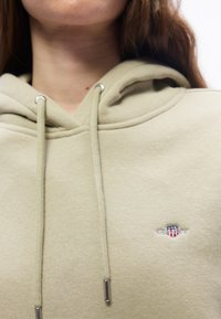 Κοντινό πλάνο ενός μπεζ φούτερ με κορδόνια και λογότυπο σε σχήμα ασπίδας με κόκκινο, λευκό και μπλε χρώμα στο στήθος.