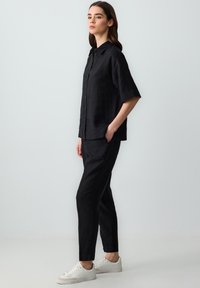 Schwarzes Leinenhemd mit kurzen Ärmeln und Knopfleiste, kombiniert mit passenden Hosen. Weiße Turnschuhe runden den Look ab.