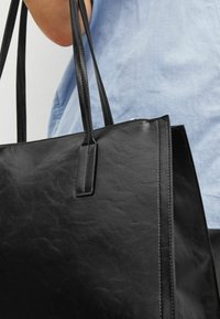 Bolso tote de cuero negro con textura suave, dos asas largas y forma estructurada. Presenta costuras mínimas y sin herrajes visibles.