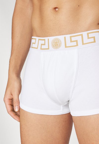 White boxer briefs made of soft fabric, featuring a gold decorative waistband with a Greek key pattern and a central emblem.