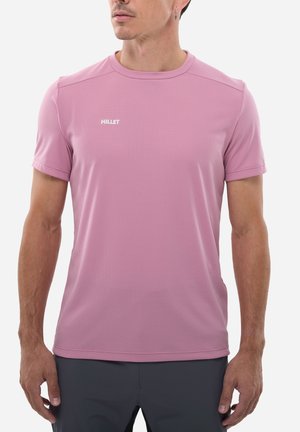 Homme portant un t-shirt de sport Millet manches courtes couleur lilas clair et un pantalon foncé, debout devant un fond uni.