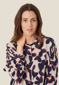 Camicia con bottoni a fiori nei toni del blu navy e arancione su uno sfondo beige chiaro. Presenta maniche lunghe e colletto rotondo.