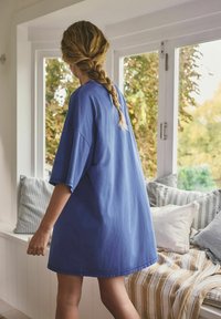 Frau mit geflochtenem Haar, die ein locker sitzendes blaues Kleid trägt und neben einem Fensterplatz mit gestreiften und einfarbigen Kissen steht.