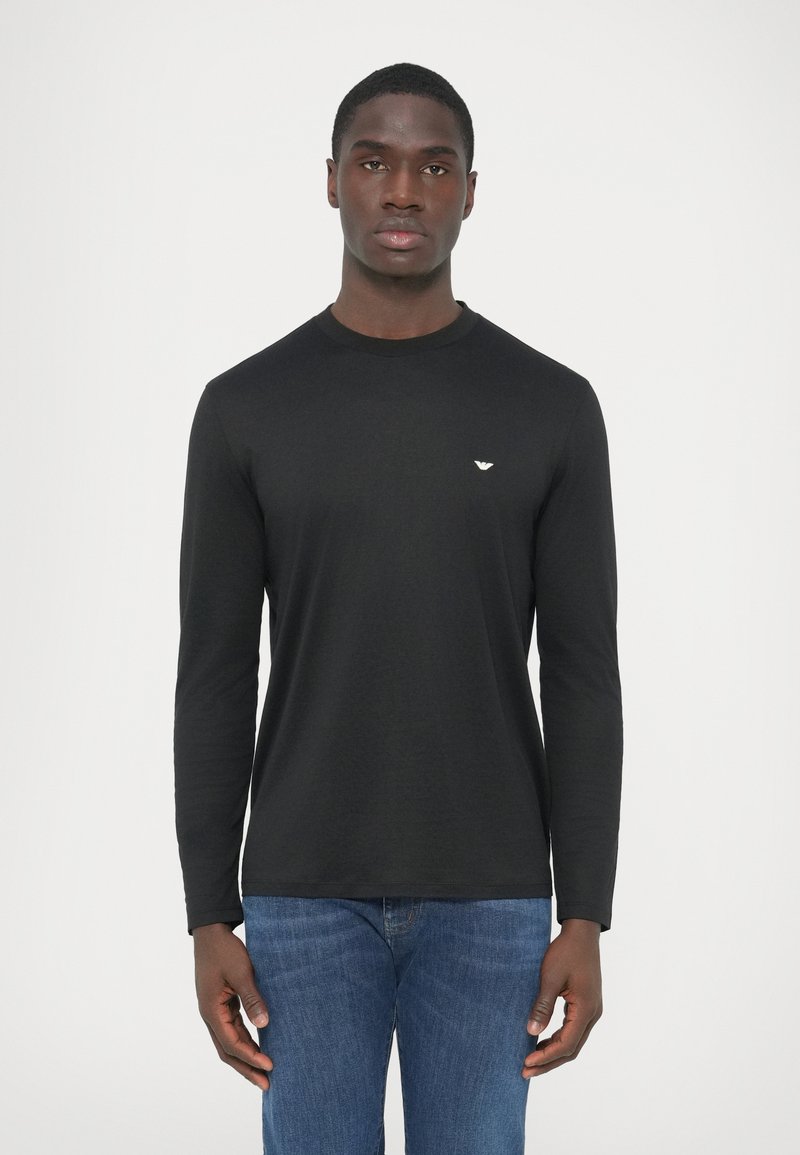 Fekete, hosszú ujjú póló puha anyagból, kerek nyakkivágással és egy kis fehér logóval a mellkason. Kék farmerral párosítva.