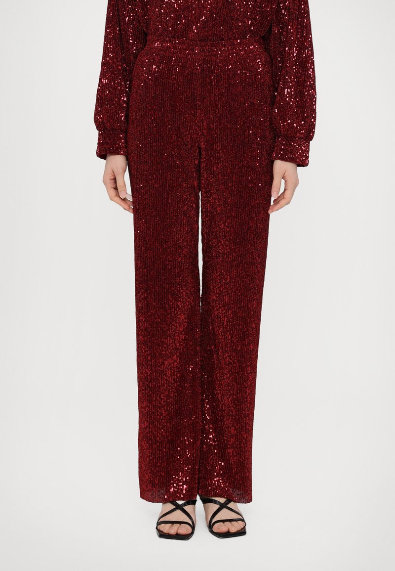Pantaloni roșii cu paiete, cu un design drept, având o suprafață texturată și un elastic la talie. Se potrivesc cu sandale negre cu curele.