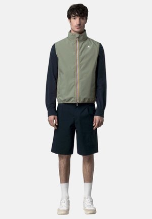 Uomo che indossa un gilet verde oliva senza maniche con cerniera, sopra una maglia nera a maniche lunghe, pantaloncini neri fino al ginocchio, calzini bianchi e scarpe da ginnastica bianche, in piedi.