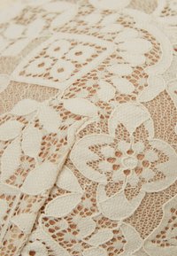 Kantstof in crème, met bloem- en bladmotieven met ingewikkelde details en een licht gestructureerd oppervlak, gelaagd over een naaktkleurige basis.