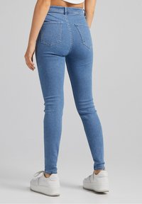 Jeans ajustados de mezclilla azul con cintura alta, que cuentan con dos bolsillos traseros y una textura de mezclilla suave, combinados con zapatillas blancas.