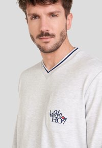 Suéter gris claro de pico con cuello a rayas azul marino, que presenta un bolsillo en el pecho con "hoHoHO!" bordado en azul y rojo. Textura suave.