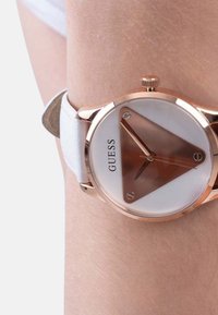 Roosa kuldne kell valge nahkrihmaga. Omab kolmnurkset pruuni ja valget kellakati, millel on minimalismile orienteeritud disain ja kolm käsi. Brändi logo on nähtav.