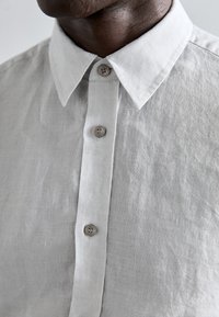 Jasnoszara lniana koszula zapinana na guziki z kołnierzykiem typu point, z dwoma guzikami na przedniej listwie oraz teksturowanym wykończeniem materiału.