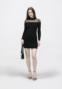 Žena v černých minisatech s dlouhými rukávy a průhledným vrškem, držící černou kabelku a obutá v černých sandálech na podpatku, stojící před bílým pozadím.