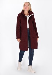 Burgundska dolga jakna iz teksturiranega blaga z visokim ovratnikom, zadrgo in stranskimi žepi. V kombinaciji z temnimi oprijetimi jeans hlačami in svetlimi čevlji.