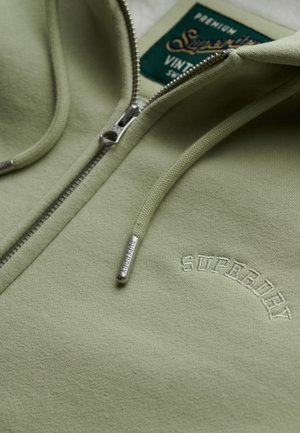 Lichtgroene zip-up hoodie met "Superdry" geborduurd op de borst, bijpassende koorden met metalen uiteinden, en een zichtbare merklabel in de kraag.