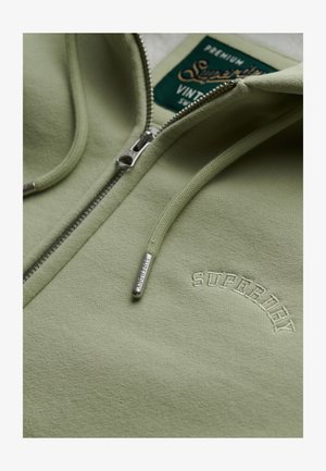 Jasnozielona bluza z zamkiem błyskawicznym z haftem "Superdry" na klatce piersiowej, dopasowane sznurki z metalowymi końcówkami oraz widoczna metka marki wewnątrz kołnierza.