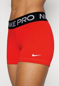 Pantalones cortos deportivos rojos con una cintura negra que presenta "NIKE PRO" en gris. Incluye un pequeño logo de Nike en blanco en la parte inferior izquierda.