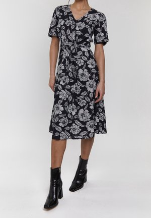 Donna che indossa un vestito midi nero con stampa floreale bianca e maniche corte, abbinato a stivaletti neri con tacco.