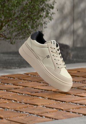 Beiger und schwarzer Low-Top-Sneaker mit dicker Sohle und Schnürsenkeln, gebrandet mit "HUGO", schwebt über einem holz- und steinmusterartigen Boden im Freien.