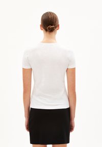 Osoba s vlasy v drdolu, oblečená v bílém tričku s krátkým rukávem a černé sukni, stojící zády k jednoduchému bílému pozadí.