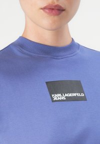 Artimas žmogaus, dėvinčio mėlyną apvalaus apykaklės džemperį su juodu kvadratiniu logotipu, kuriame užrašyta "KARL LAGERFELD JEANS" ant krūtinės.