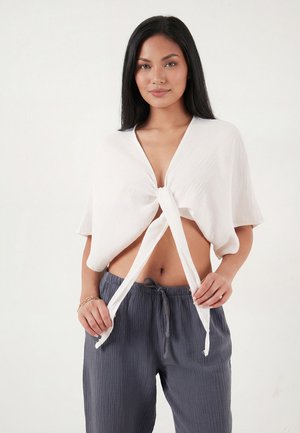 Vrouw met lang zwart haar, een wit T-shirt met korte mouwen dat in de taille is gestrikt, en losse donkergrijze broek met trekkoord.