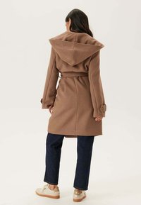 Femme portant un manteau capuche ceinturé marron, un pantalon bleu foncé et des baskets beige, se tenant de dos contre un fond clair.