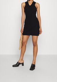 Robe sans manches en maille côtelée noire avec un décolleté en V, coupe ajustée. Associée à des mules à talons blocs noires et à un sac tressé beige.