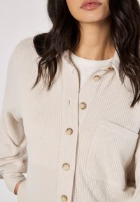 Beige button-up shirt with a textured ribbed pattern, featuring a chest pocket and light-colored buttons. Soft fabric, relaxed fit.