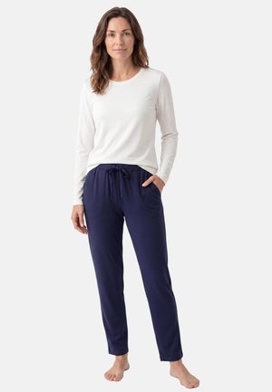 Femme debout pieds nus portant un haut blanc à manches longues et un pantalon bleu marine à cordon, sur un fond clair uni.