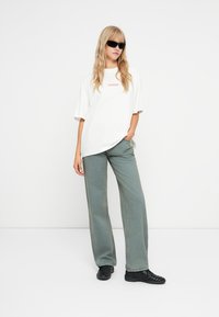 Λευκή oversized μπλούζα με κείμενο "PEGADOR", σε συνδυασμό με χαλαρές γκρι-πράσινες τζιν παντελόνες και μαύρα αθλητικά παπούτσια; το μοντέλο ποζάρει με γυαλιά ηλίου.