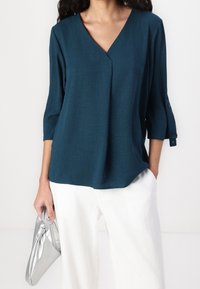 Teal, gestructureerde blouse met een V-hals en driekwartmouwen. Gecombineerd met witte, brede broekspijpen en een zilveren, geknoopte clutch.