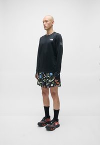 Svart langermet skjorte med teksturert stoff, kombinert med flerfargede shorts med blomstermønster og svarte joggesko. Svarte knesokker.
