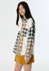 Camisa de botonadura con una mezcla de tela blanca texturizada y patrones de cuadros en azul oscuro, beige y marrón. Presenta un bolsillo en el pecho y puños elásticos.