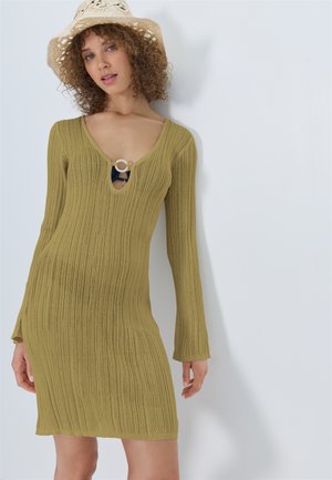 Femme aux cheveux bouclés portant un chapeau de paille et une robe côtelée vert olive avec des manches longues et un détail d'encolure circulaire.