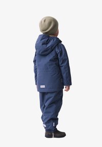 Marine waterdichte jas met capuchon, elastische manchetten, gedragen met bijpassende broek en bruine schoenen. Lichtgroene gebreide beanie op het hoofd.