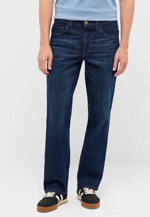 Mann trägt dunkelblaue Jeans mit geradem Bein, schwarz-weiße Sneakers und ein hellblaues T-Shirt vor einem einfarbigen Hintergrund.