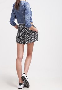 Žena stojí s rukama v kapsách, oblečená v zkrácené džínsové bundě, černo-bílých květovaných šortkách a bílých teniskách na neutrálním pozadí.