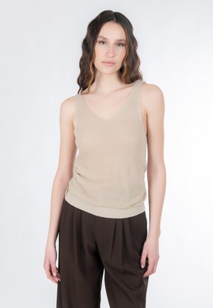 Femme aux longs cheveux ondulés portant un haut tricoté sans manches beige et un pantalon brun foncé taille haute, sur un fond blanc uni.