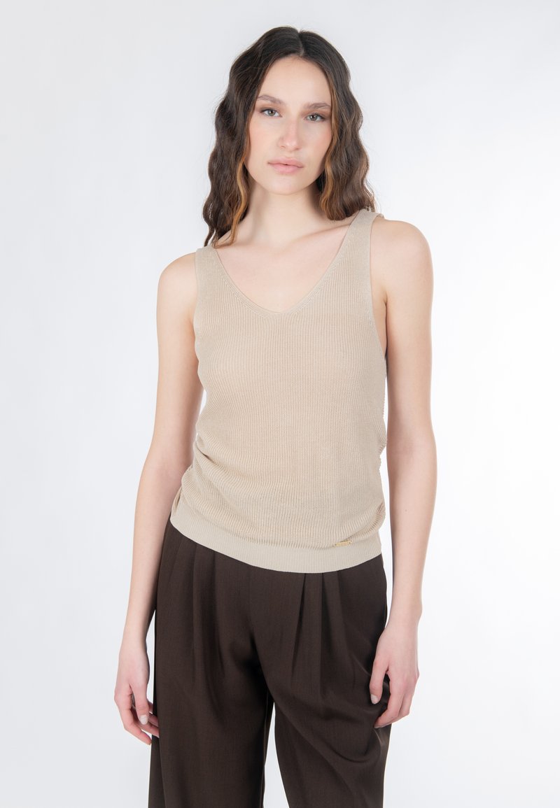 Mujer con cabello largo y ondulado que lleva un top de punto sin mangas beige y pantalones de talle alto marrón oscuro, sobre un fondo blanco liso.