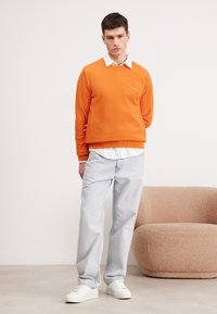Oranje textuurtrui met een ronde halslijn, gelaagd over een witte blouse met kraag, gecombineerd met lichtblauwe jeans en witte sneakers.