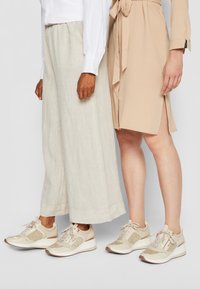 Beige sportieve schoenen met een gestructureerde afwerking, glinsterende accenten en gelaagde zolen. Draagt met lichte linnen broek en een beige jurk.