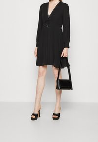 Vestito nero plissettato con scollatura a V profonda, maniche lunghe e dettaglio con fiocco in vita. Abbinato a una borsa nera e tacchi a piattaforma.