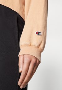Camicetta a maniche lunghe in cotone beige chiaro con polsini a coste. Presenta un piccolo logo ricamato sulla manica sinistra. Abbinata a tessuto nero.