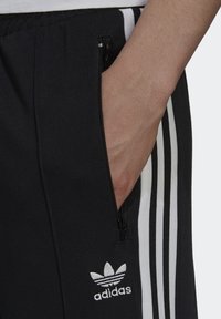 Černé sportovní kalhoty slim fit s bílými bočními pruhy, kapsou na zip a bílým logem na dolní části nohavice. Hladký textilní povrch.