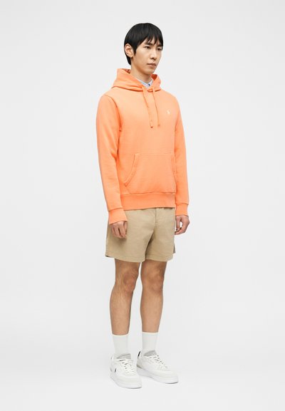 Νέος άνδρας που στέκεται, φοράει πορτοκαλί φούτερ με κουκούλα, μπεζ σορτς, λευκές κάλτσες και λευκά αθλητικά παπούτσια σε απλό ανοιχτόχρωμο φόντο.