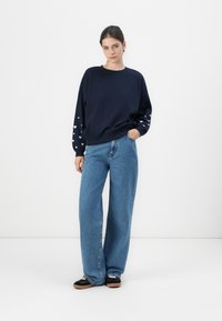 Marineblå genser med hvite hjerte mønstre på ermene, kombinert med lys blå vidde jeans og svarte sneakers med brune såler.