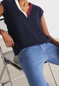 Top senza maniche di colore blu navy con scollo a V, caratterizzato da dettagli bianchi e granata; abbinato a jeans azzurri chiari, su una sedia in metallo cromato. Texture liscia.