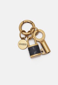 Zlatý klíčový řetízek s černým mini zámkem, klíčem a kulatým štítkem s vyrytým nápisem "GUESS." Texturovaný povrch zámku a kulatý design kroužku na klíče.