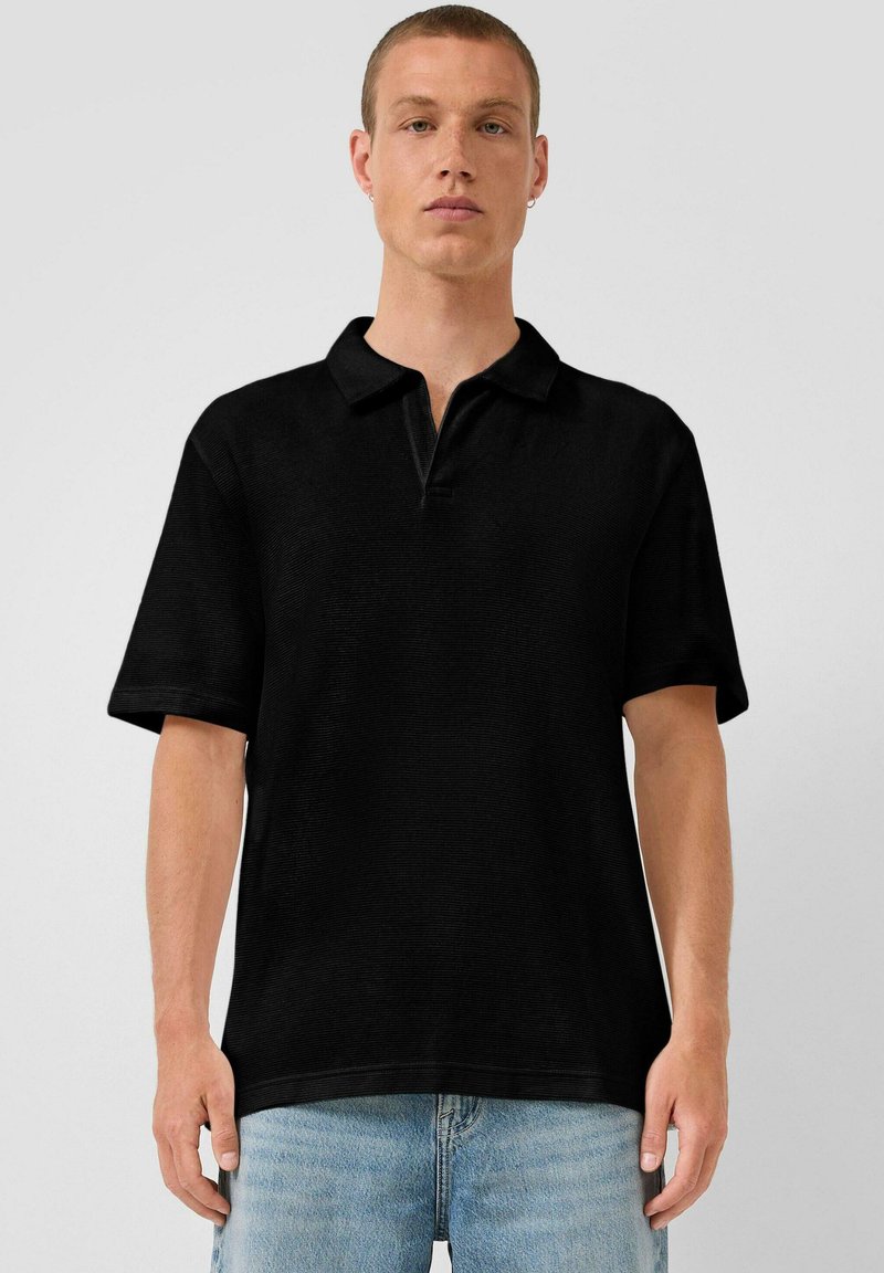 Junger Mann, der ein schlichtes schwarzes Poloshirt mit kurzen Ärmeln und hellblaue Jeans trägt, vor einem schlichten hellgrauen Hintergrund steht.