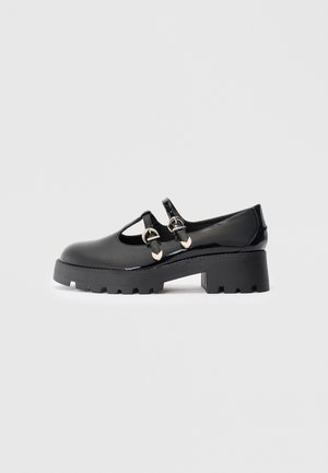 Black patent leather Mary Jane shoe with chunky sole and two silver buckle straps, side view on neutral background.